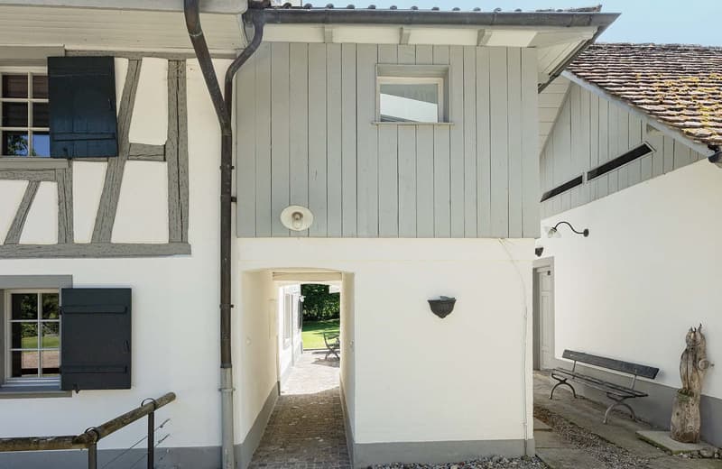 Two-story house with a wooden exterior, white walls, and a tiled roof. The house has a covered entryway leading to a garden or outdoor area. There is a bench or seating area visible outside the house.