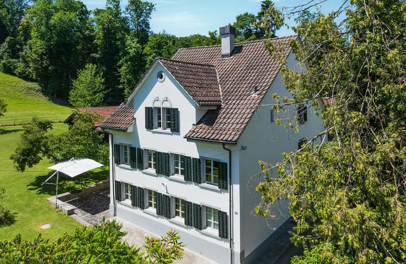 Two-story white house with red tile roof, black shutters, and a balcony or terrace. The house is surrounded by lush greenery and trees.