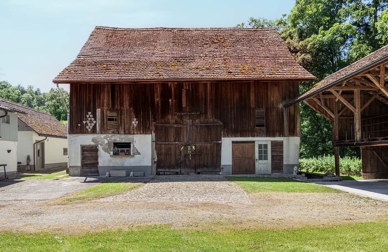 A large, rustic wooden barn with a red tile roof, surrounded by lush greenery and trees. The barn has a central wooden door and several smaller windows and doors along the sides. The building appears to be well-maintained and situated in a picturesque rur