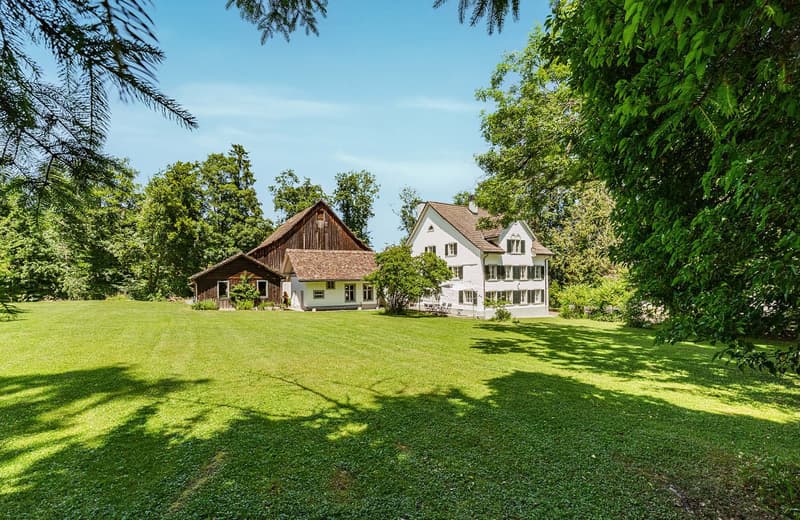 Two-story white house with a wooden barn-like structure, surrounded by lush green trees and a well-manicured lawn. The property appears to have a balcony or terrace.