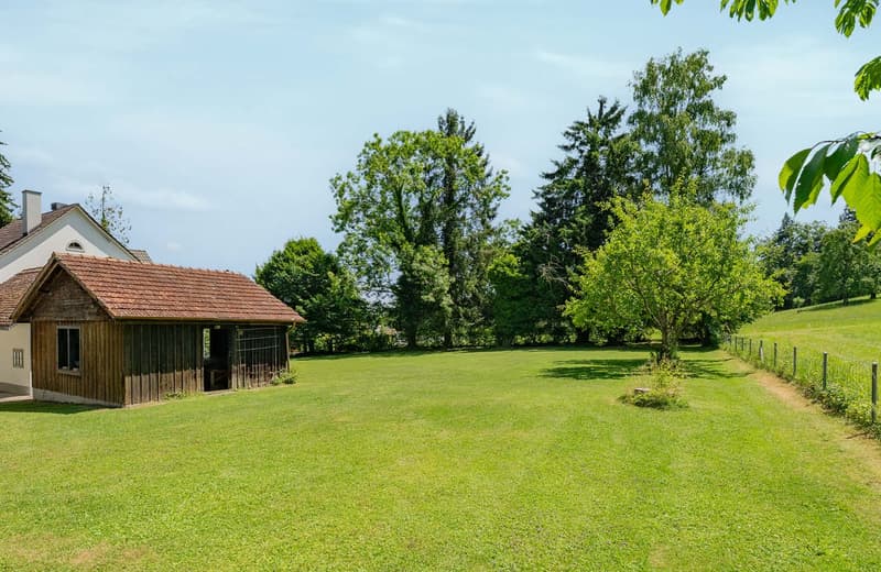 A wooden cabin-style house surrounded by a large grassy lawn, with trees and greenery in the background. The house has a covered porch or balcony area.