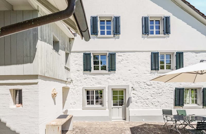 2-story white stone house with black shutters, wooden beams, and a patio area with outdoor furniture