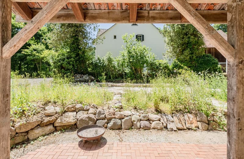 Rustic wooden structure with a tiled roof, surrounded by lush greenery and a stone wall. There is a brick patio area with a fire pit or grill in the foreground.