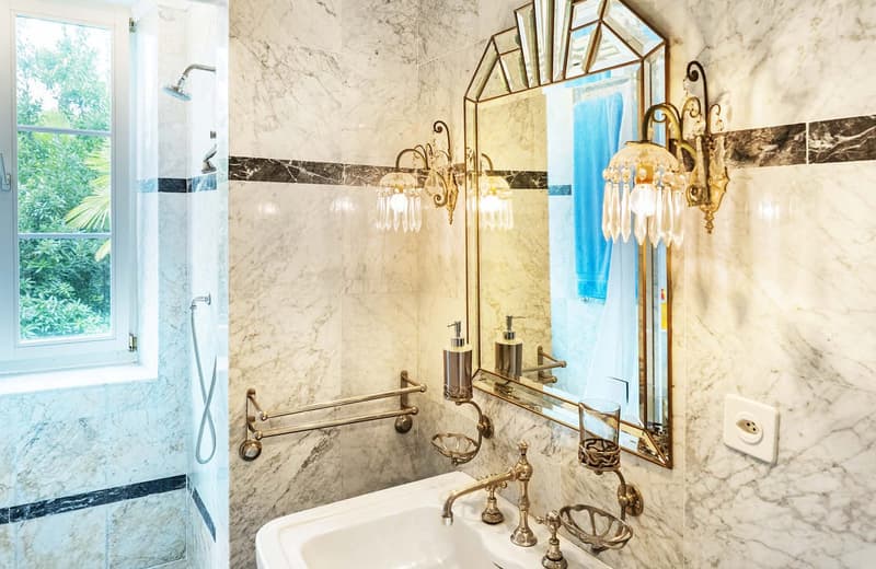 Bathroom with white marble walls, brass shower head, vanity with golden faucet, mirror with Art Deco style wall lamps, power outlet.