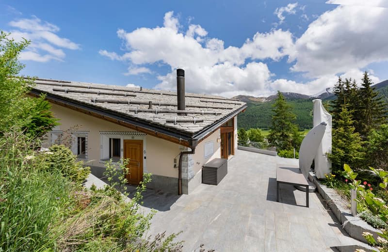 2-story mountain chalet with a stone and wood exterior, a slanted roof with a chimney, and a paved terrace overlooking a forested mountain landscape