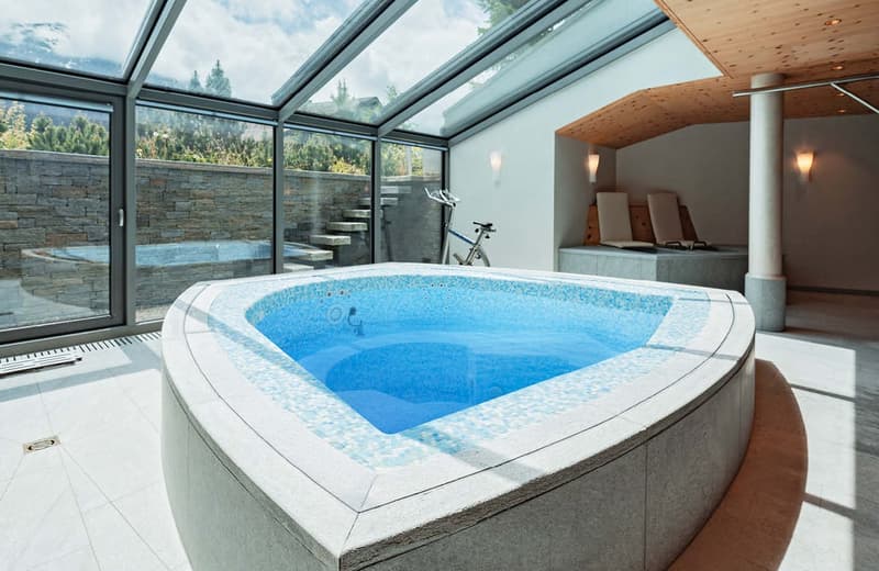 Large indoor hot tub or spa with glass walls and ceiling, surrounded by stone walls and wooden accents, with a view of the outdoor forest landscape
