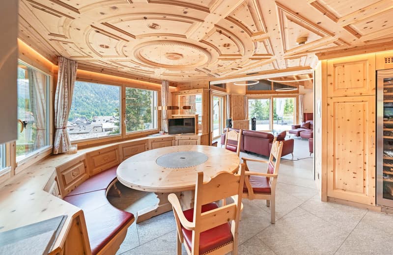 Spacious living room with large windows offering a scenic mountain view, ornate wooden ceiling with decorative moldings, round wooden dining table, red leather chairs, and a cozy seating area with sofas.