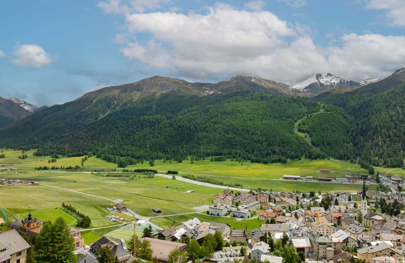 The image shows a picturesque mountain landscape with a small town nestled in the valley below. The town is surrounded by lush green forests and meadows, with snow-capped peaks in the distance. The scene features a mix of residential and commercial buildi