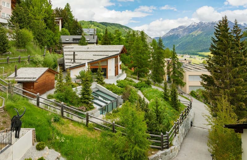 The image shows a scenic mountain landscape with a village nestled among the trees. The buildings have a traditional alpine style with wooden and stone construction. There are walking paths and fences winding through the lush, green vegetation. In the bac