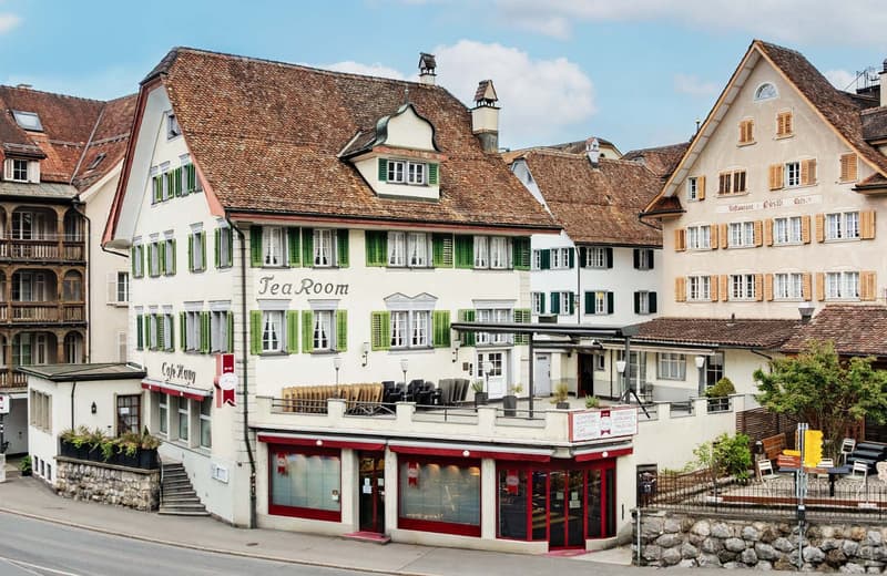 A multi-story building with a traditional European architectural style, featuring red-tiled roofs, green shuttered windows, and a balcony. The building houses a tea room on the ground floor, with outdoor seating. The surrounding buildings have a similar s