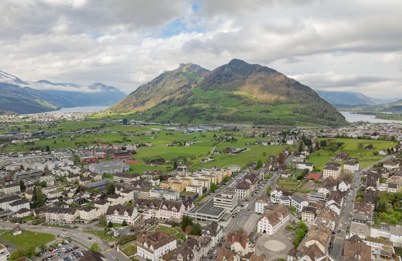 Aerial view of a mountainous town with a mix of residential and commercial buildings, surrounded by lush green hills and mountains. The town appears to be situated near a body of water, possibly a lake or river. The scene depicts a picturesque and scenic