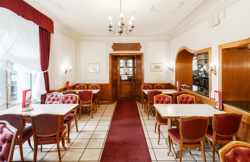 This image shows the interior of a traditional European-style cafe or restaurant. The space features a warm, cozy atmosphere with wooden furniture, including tufted red leather chairs and tables, as well as a decorative wooden archway and chandelier. The