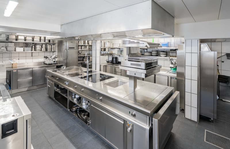 Large commercial kitchen with stainless steel appliances, cooking equipment, and storage shelves. The kitchen appears to be well-equipped for a professional food service operation.