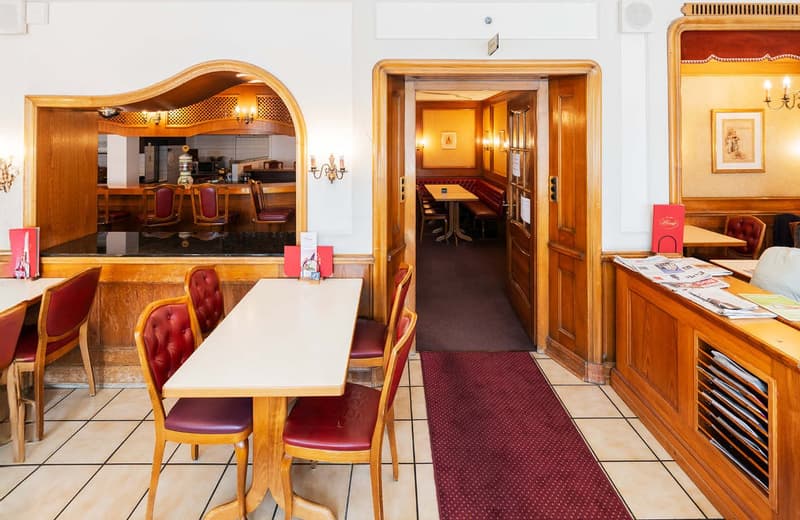 The image shows the interior of a cozy restaurant or cafe. The space features warm, wooden decor with arched doorways and a fireplace visible in the background. There are several tables with red leather chairs, and a bar area can be seen through an openin