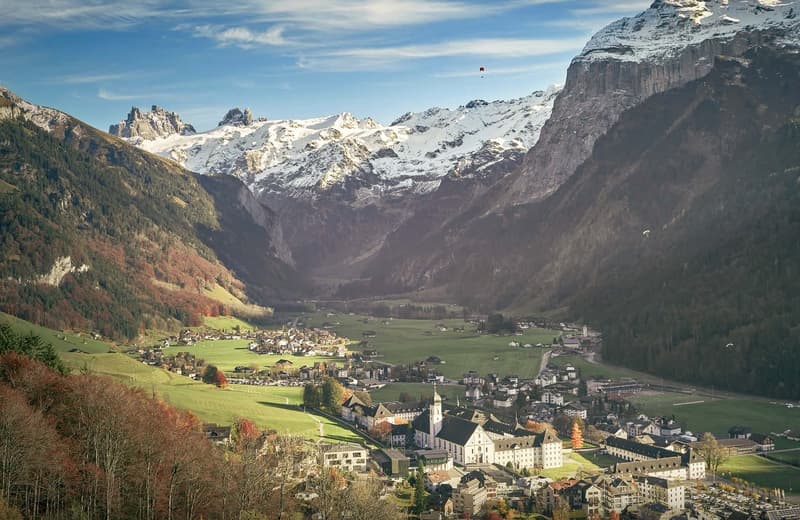 Mountain village with snow-covered mountains, a large church, and a clear blue sky with clouds.