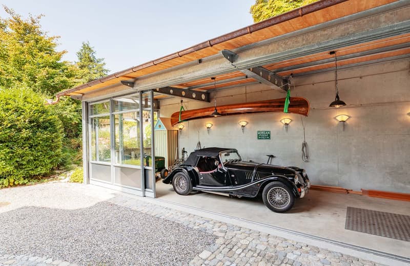 Classic vintage car parked inside a modern garage with glass walls, a concrete floor, and a roof with lighting fixtures. A canopy roof covers the entrance, with trees and bushes nearby.