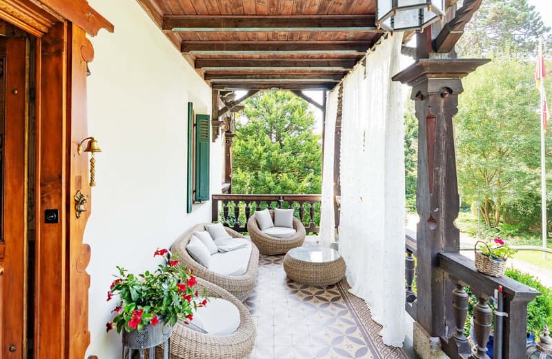 Terrace with wooden floor, surrounded by wicker chairs, central table, white walls, white curtains, green shutters, and plants