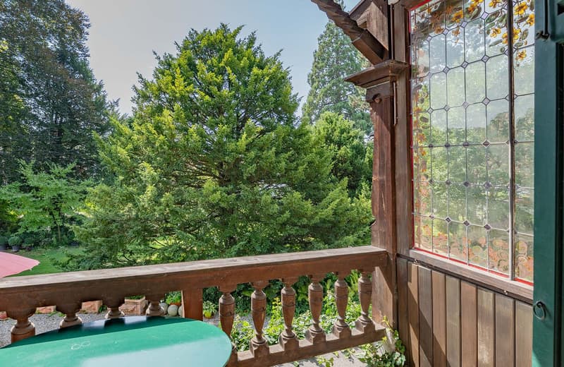 Balcony with wooden railing, green round table, potted plants, view of trees, and colorful glass window