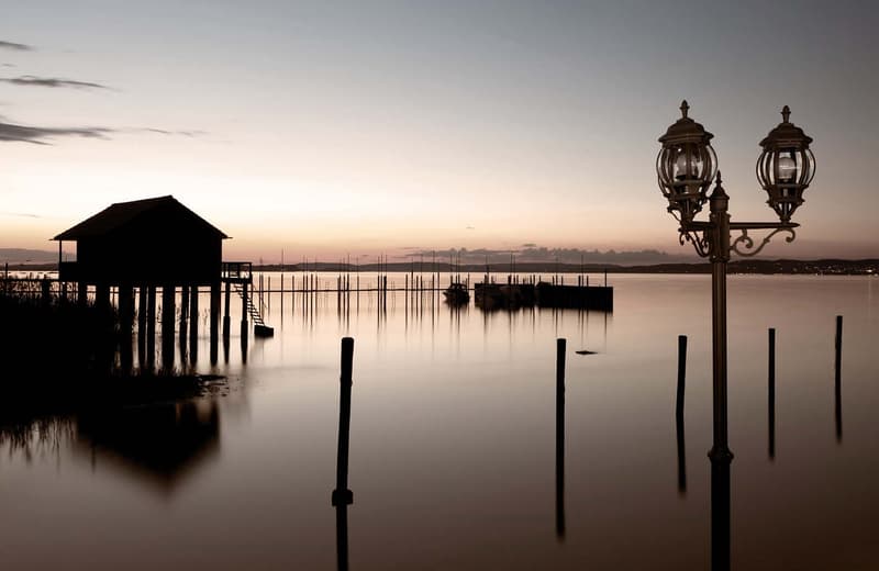 The image shows a serene lakeside or coastal scene at sunset. There is a wooden dock or pier extending into the calm, reflective water, with several wooden posts or pilings visible. In the background, there is a small building or structure silhouetted aga