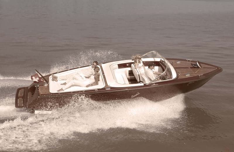 A vintage-style wooden speedboat speeding across the water, with people seated inside the boat.