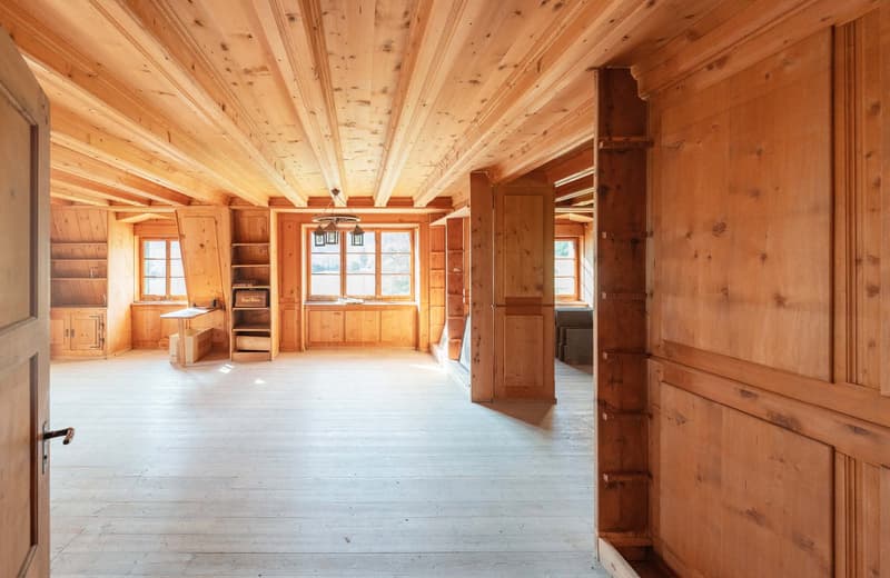 This image shows a large, open-plan room with wooden walls and ceilings. The room has a rustic, cabin-like feel, with exposed wooden beams and paneling. There are several windows that let in natural light, and the floor appears to be made of wood planks.