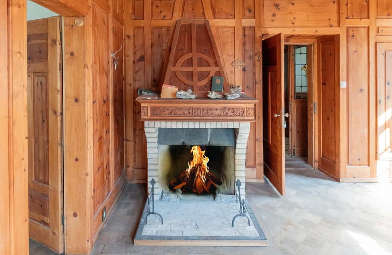 Rustic wooden interior with a large stone fireplace, ornate wooden mantelpiece, and a cozy fire burning inside