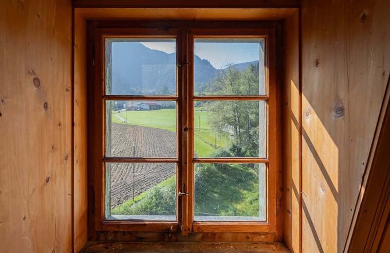 The image shows a wooden window frame with a scenic view of a rural landscape. The window overlooks a green field or meadow, with mountains and trees visible in the distance. The wooden walls of the room provide a rustic, cozy atmosphere.