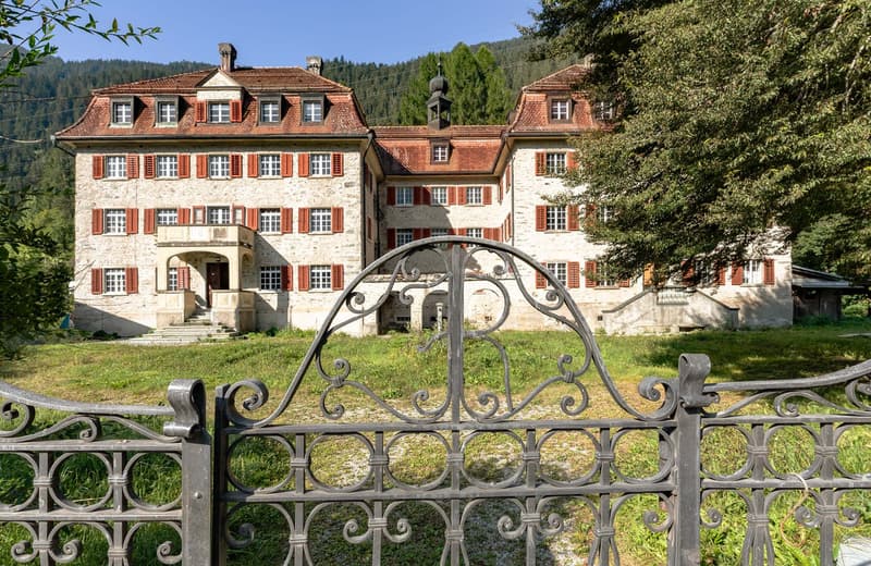A large, historic stone building with red-tiled roofs and ornate windows. The building has a central tower and is surrounded by lush greenery. There is an ornate wrought-iron gate in the foreground.