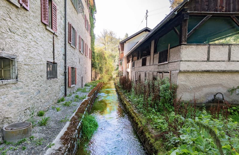 The image shows a picturesque alleyway in a historic European town. The alleyway features a small stream or canal running through it, with stone walls and wooden buildings on either side. The buildings have traditional shuttered windows and some have balc