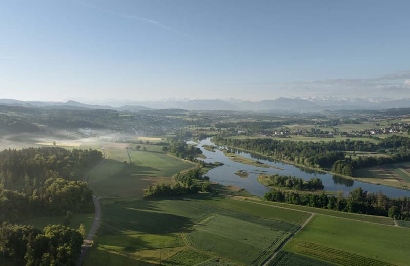 Aerial view of a scenic rural landscape with rolling hills, green fields, a winding river, and distant mountains in the background.