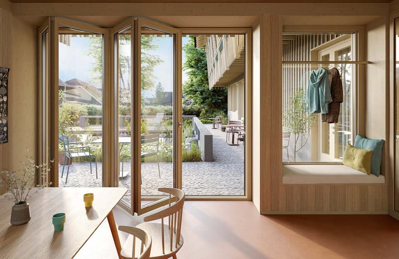 Spacious living room with large windows overlooking a patio and garden area, wooden furniture including a round dining table and chairs, and a built-in window seat with cushions