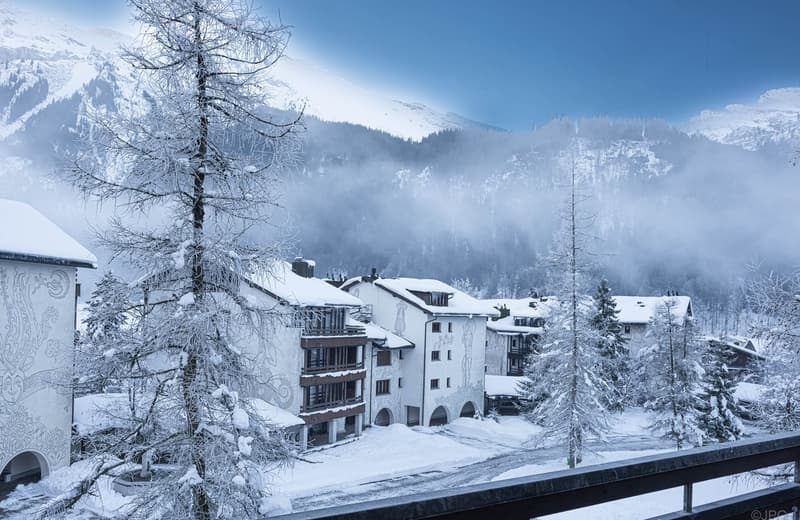 cluster of apartment buildings, snow-covered trees, snowy mountains, snow on rooftops