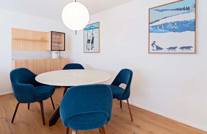 Dining area with round white marble table and blue cushioned chairs. Wooden cabinet, counter top, and a coffee maker.
