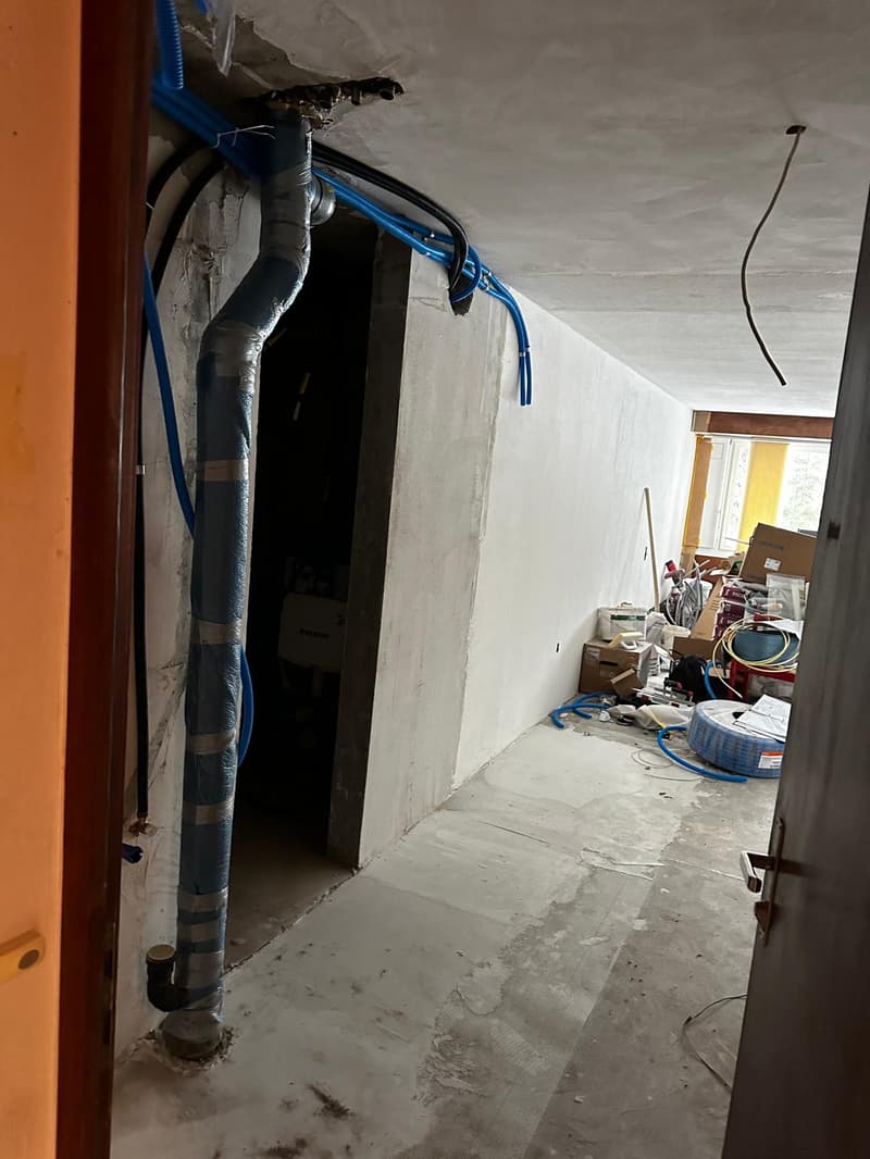 Concrete walls, grey tiled floor, exposed pipes and cables, storage boxes and tools scattered around.