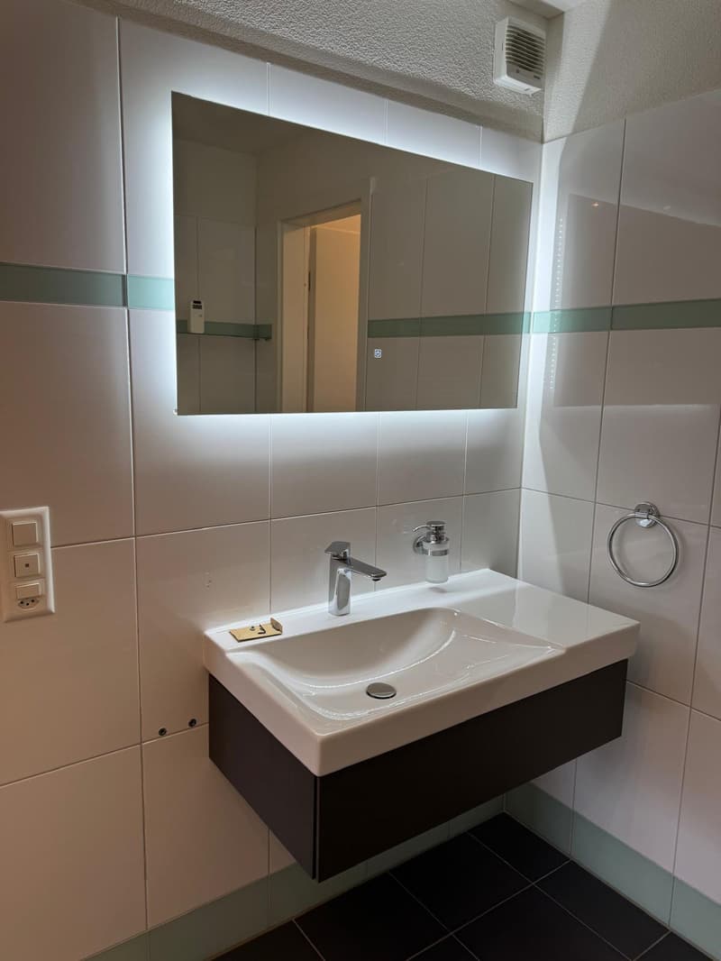 modern bathroom with white tiles, mirror with lighting, sink with faucet, soap dispenser, towel ring, power outlet