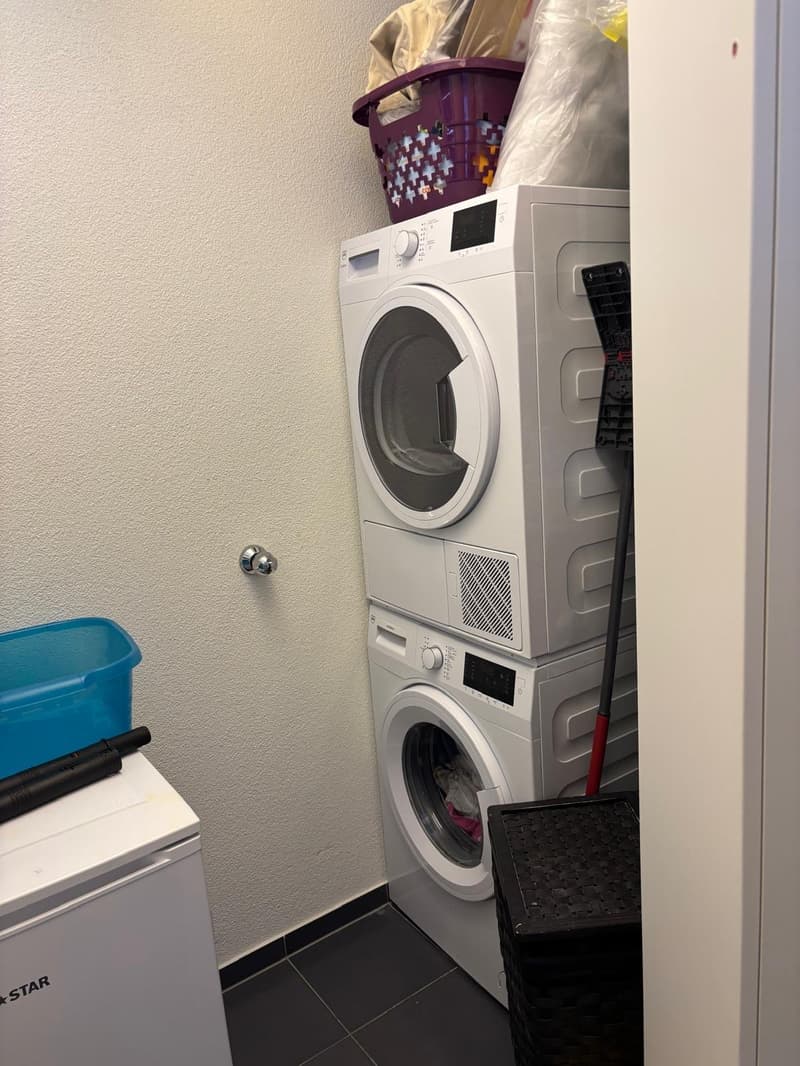 Laundry room with stacked washing machine and dryer, storage boxes, and a basket on top of the dryer