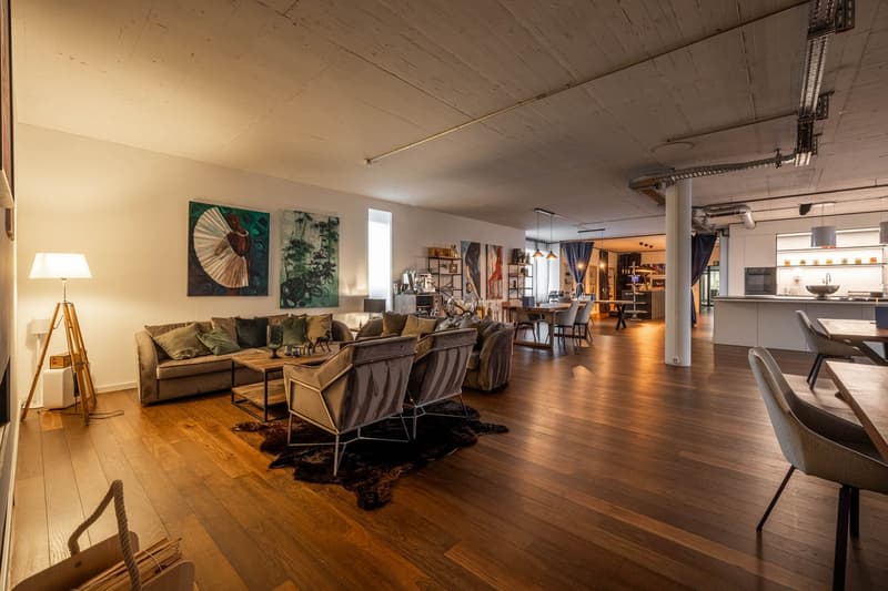 Open living space with wooden floors, large couch, coffee table, dining table, chairs, lamps, paintings, kitchen area