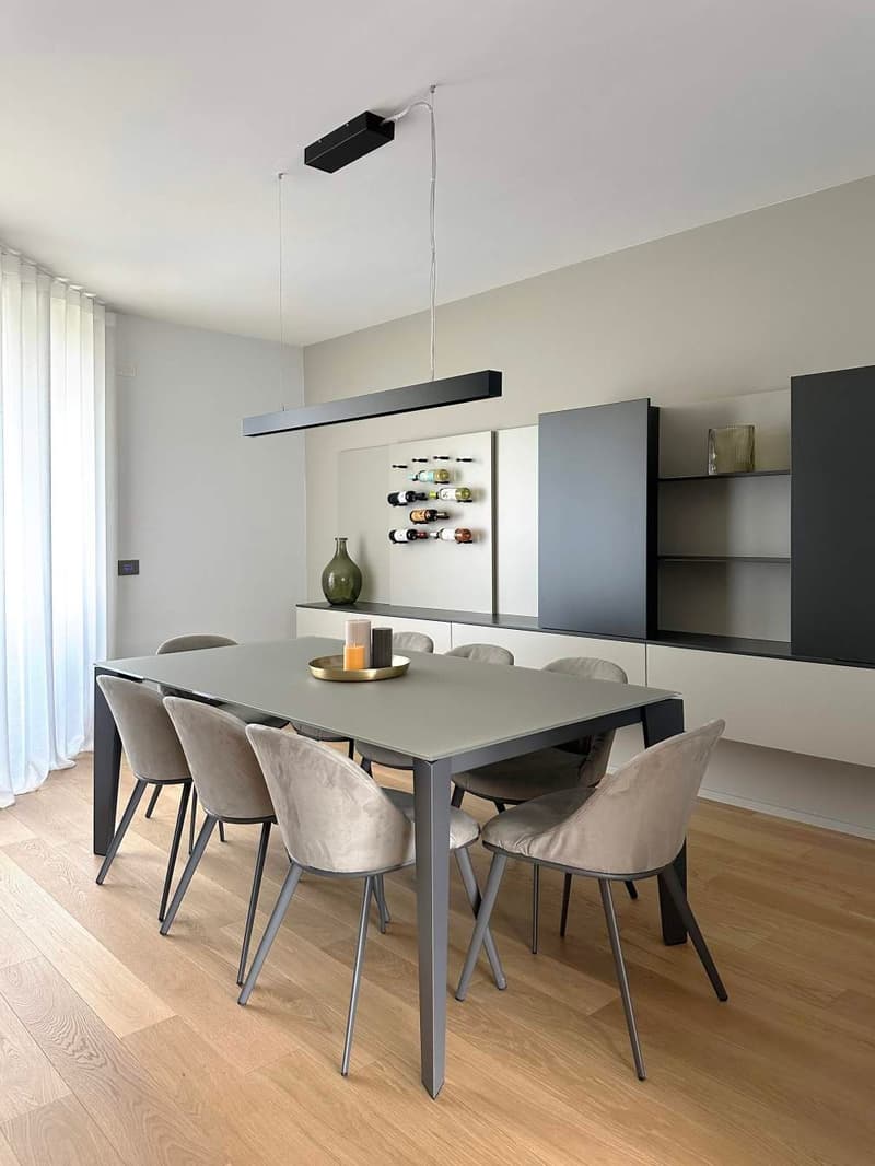 Rectangular grey dining table with six modern grey chairs. Table has wooden top and metal legs. Grey shelving units with shelves on the wall behind the table. Large wine rack with several bottles. Overhead ceiling lighting. Grey marble tile floor.