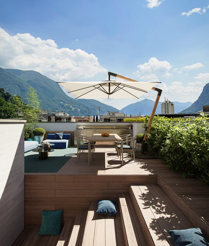 wooden deck, dining table with parasol, blue couch, pillows, small table, bar, potted plants, mountain view in the background