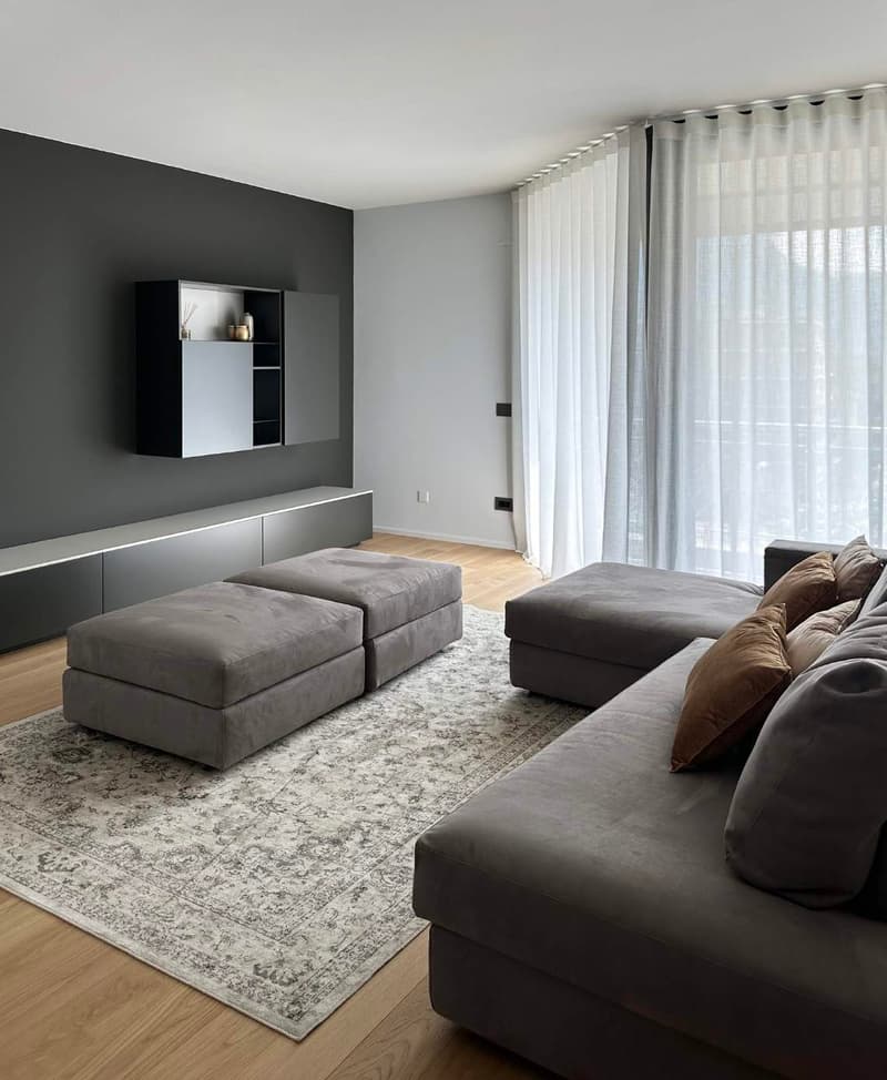 modern living room, gray walls, large gray sofa, two gray ottomans, gray rug, gray wall cabinets, television mounted on the wall, white curtains