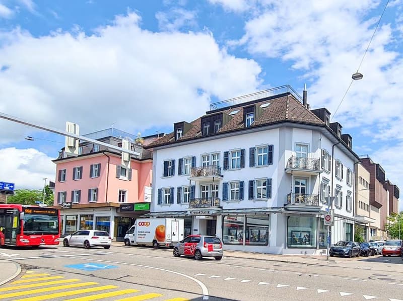 Three-story residential building with shops on the ground floor, balcony, and parked cars on the street.