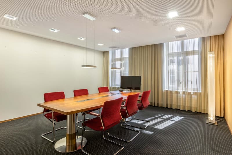 Long wooden table, red chairs, monitor on table, beige curtains, white walls, carpeted floor