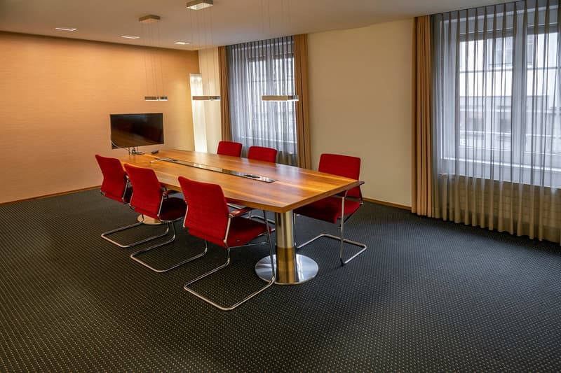 Meeting room with wooden desk, red chairs, flat screen TV, two windows with curtains, wooden floor, beige walls