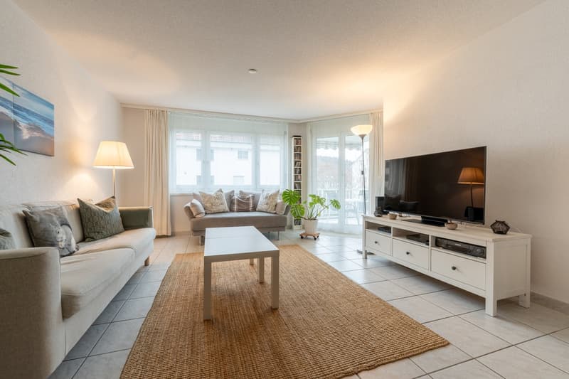 Neutrally decorated living room with a sofa, coffee table, entertainment center, and a flat-screen TV. There are large windows with white curtains, a shelf with books, and a plant. A carpet and lamps provide additional comfort.