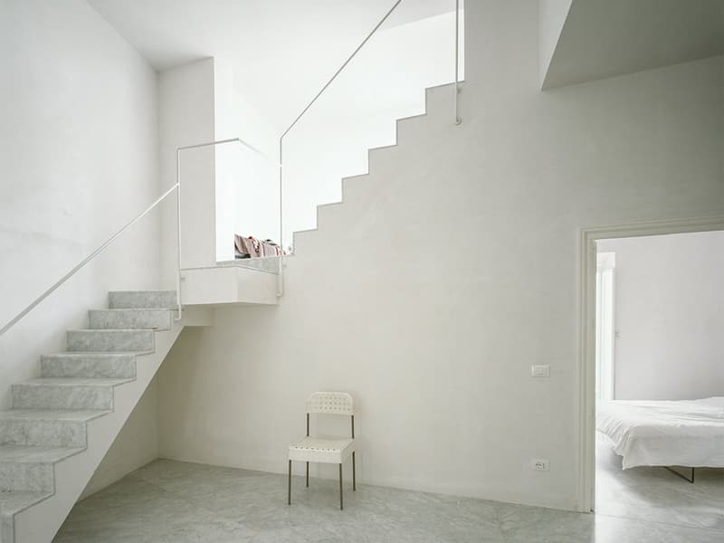 White marble staircase, white walls, open doorway leading to bedroom with a bed, small white chair, window with pillows on window sill.