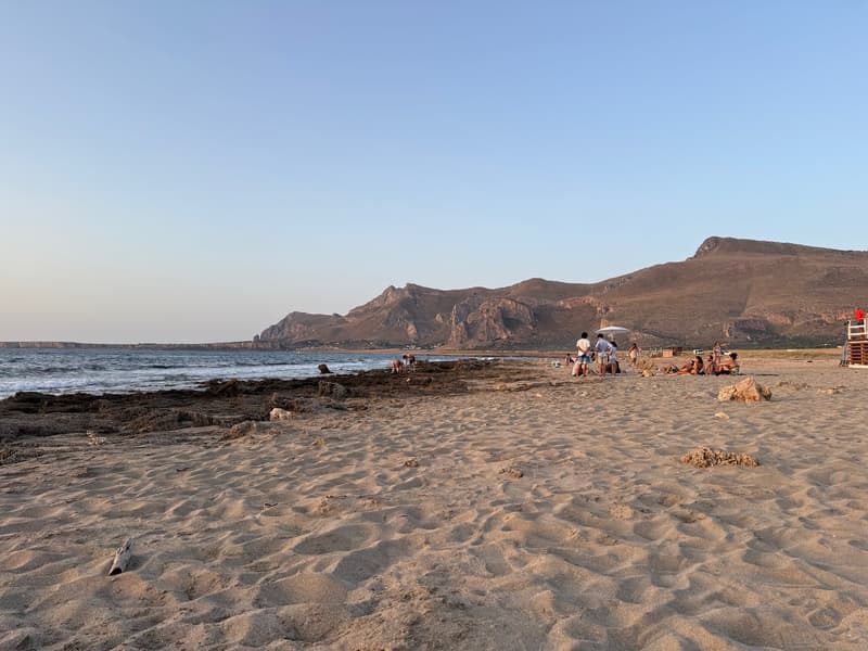 Beach with sand, mountains in the background, clear sky, few people sitting and standing on the beach