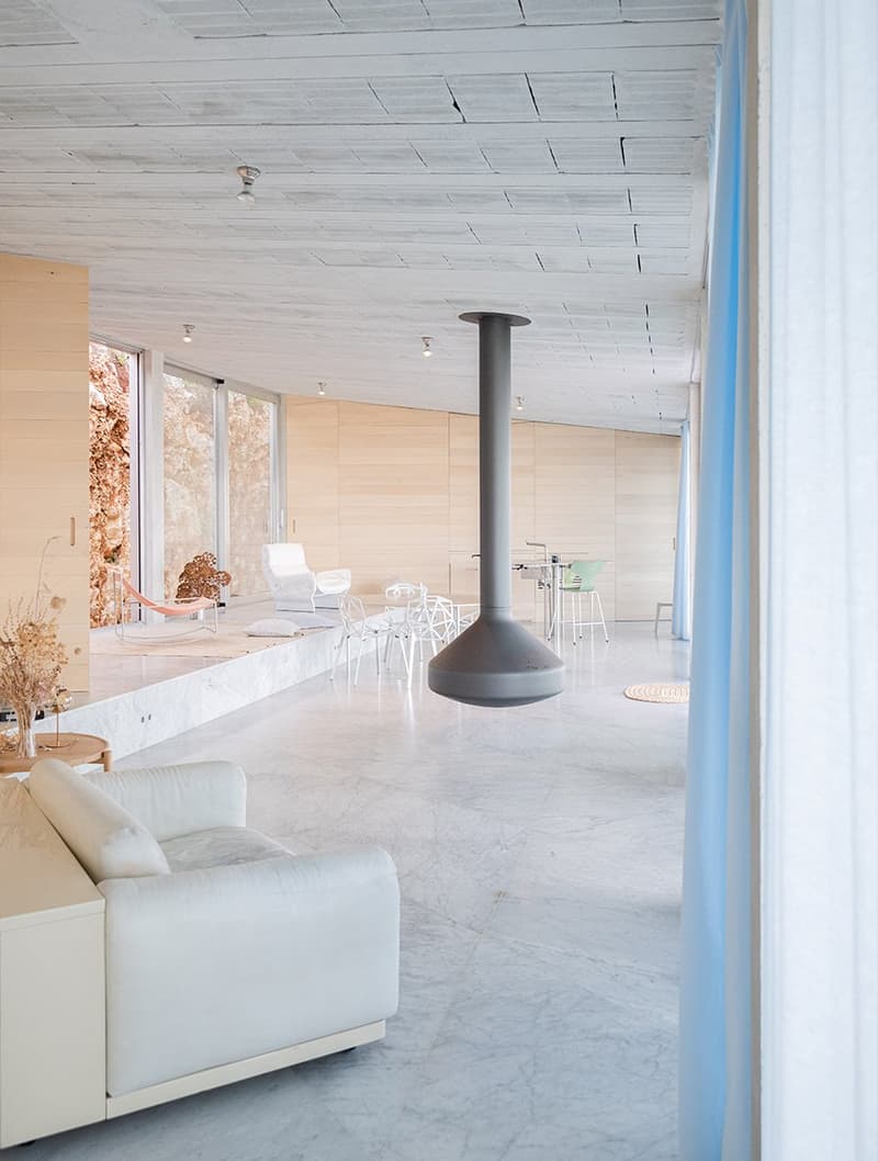 Minimalist interior with large white couches, large glass windows, white marble flooring, hanging cylindrical fireplace, white walls, white chairs, white dining table, dried plants