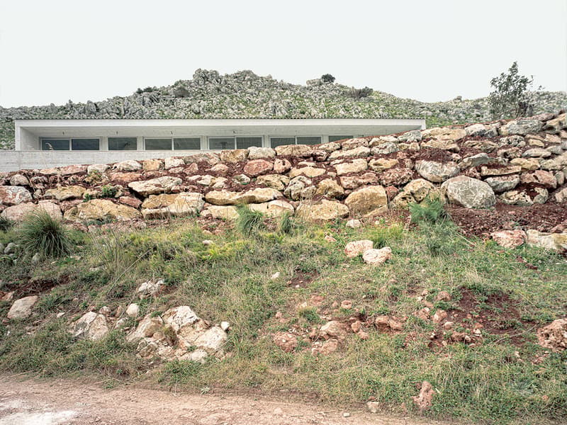 Modern architectural design, white walls, large rock wall in front, numerous windows, grass and small plants on the hill in the background, elevated ground, view of a mountain range
