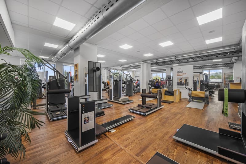 Well-equipped gym with a wooden floor, multiple exercise machines, and large windows allowing natural light.
