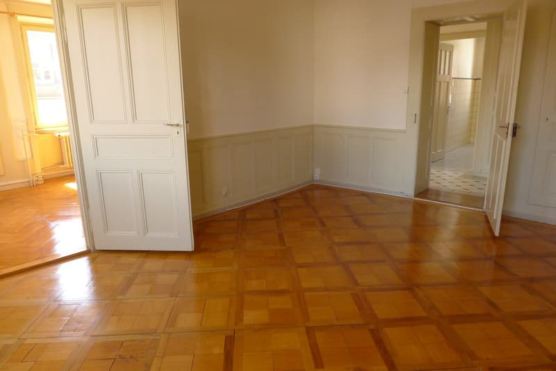 Wooden floor, white walls, two open doors, radiator, electrical outlets, hallway
